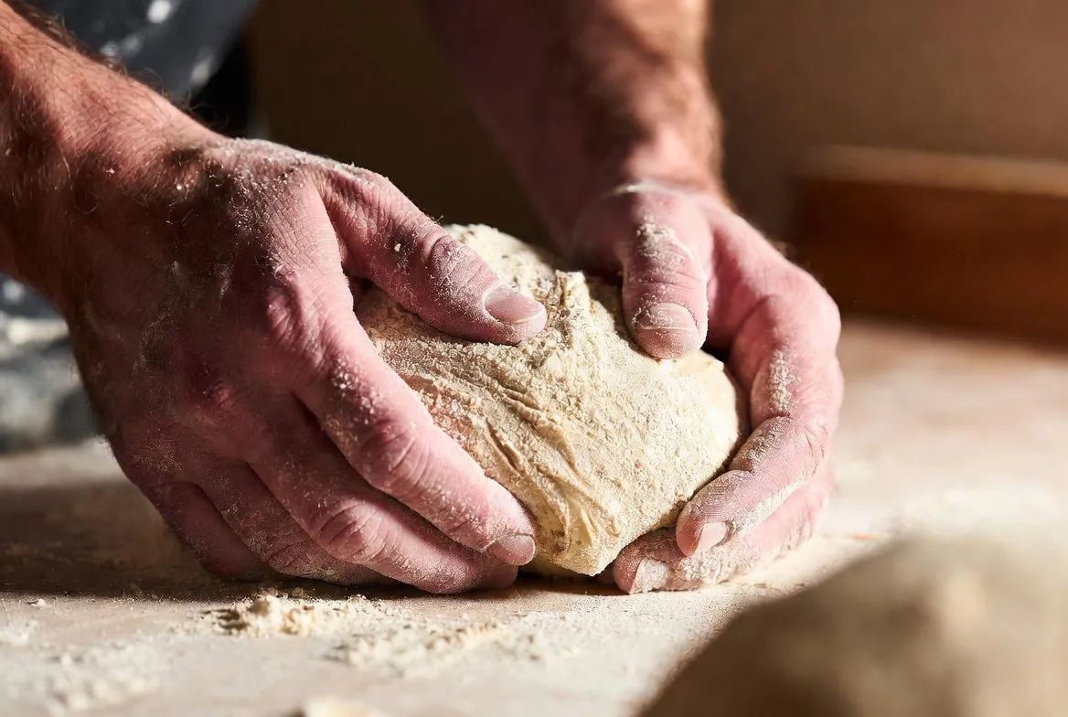 Baker’s flour-covered hands shaping fresh sourdough dough on a wooden bench